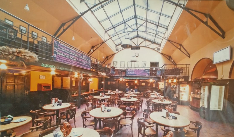 prague beer halls u vejvodu interior. 14 round tables each with 5 wooden chairs. a balcony on the left side looking up to a roof light running the length of the ceiling