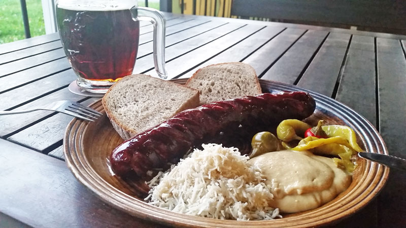 grilled czech sausage on a plate with slices of bread, horseradish, czech mustard and peppers. The plate is on a table and there is a glass of beer next to it