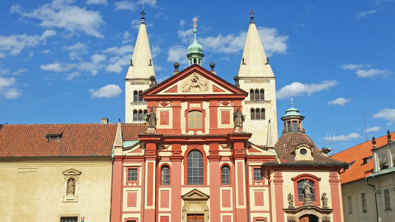 prague castle romanesque design st george basilica in light red colour with two asymmetrical white gothic towers behind it and a blue sky with light clouds