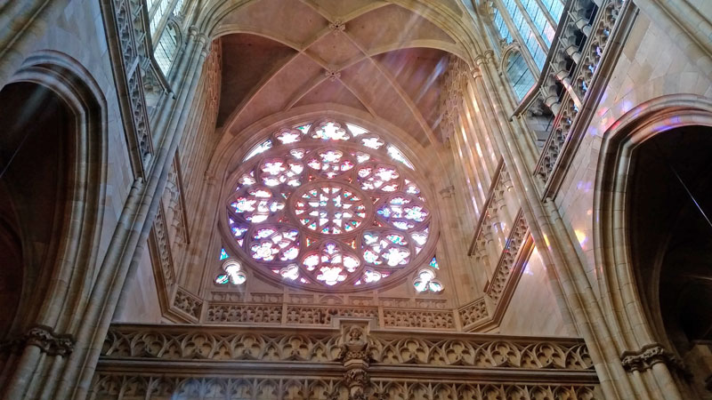 The Prague Castle Saint Vitus West Entry Rose Window viewed from inside with visible coloured light beams and reflections off of the stone walls