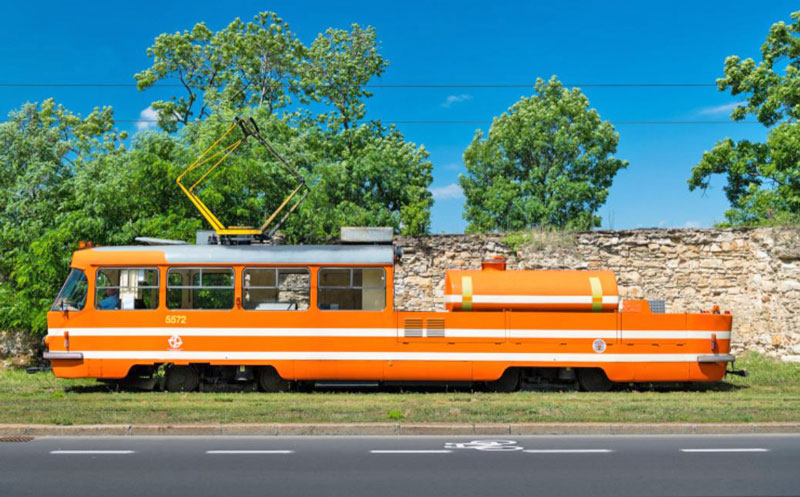 the prague lubrication tram, orange with white stripes, a rough stone wall behind, trees and blue sky