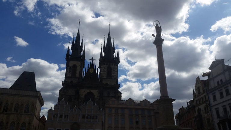 silhouette against a blue sky with white clouds of the prague marian column and the tyn church