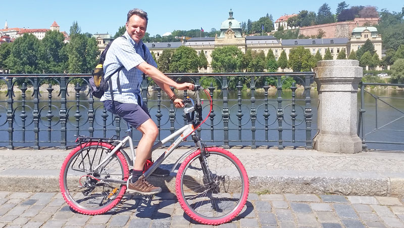 man on bike with red tyres with river behind and the prague city hall building