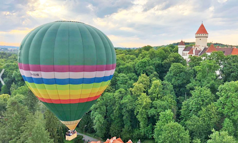 rainbow coloured prague hot air balloon over a forest and passing a castle at low level