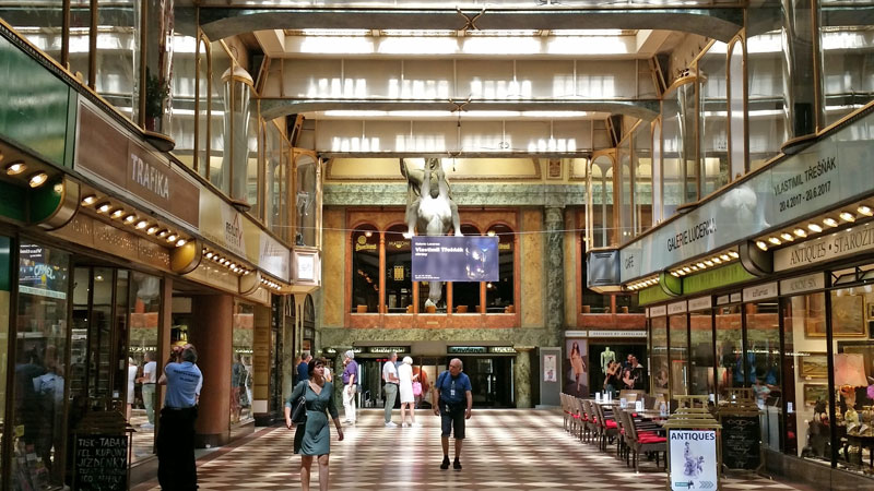 the 1920's prague lucerna passage viewed from one end with chequered tile floor, glass fronted shops on both sides. at the end are 5 tall roman arched windows with marble surround. in front is a sculpture of a king sitting on a horse which is upside down with it's feet tied and hanging from the ceiling