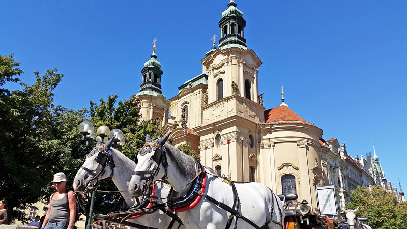 twin towers of the baroque church of st nicholas in prague old town with horses and carriage and blue sky