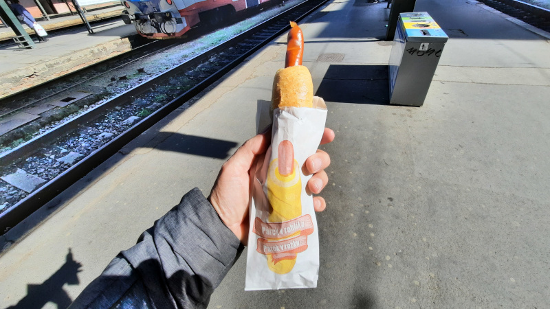 man on a railway station platform holding a czech hot dog called parek